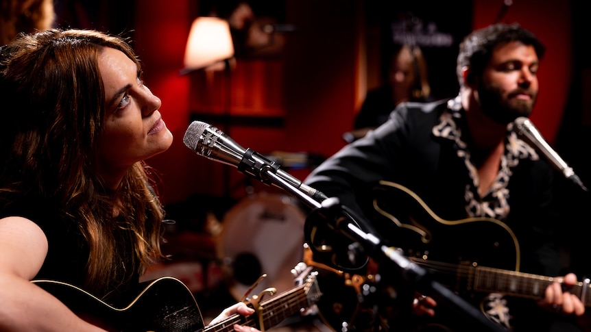 Woman on the left and man on the right, both sitting down holding guitars and have microphones in a studio