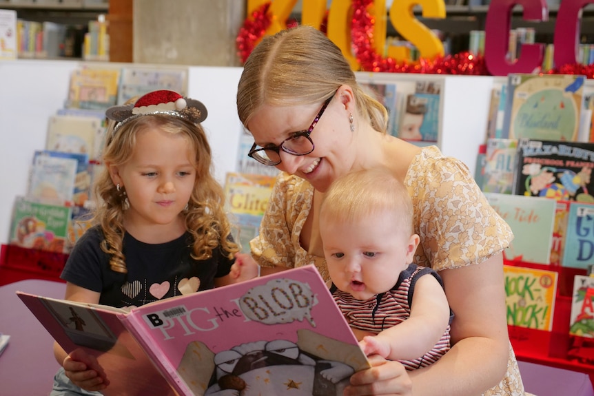 Mum sitting with young child and baby, holding a picture book in front of her and reading to them. 