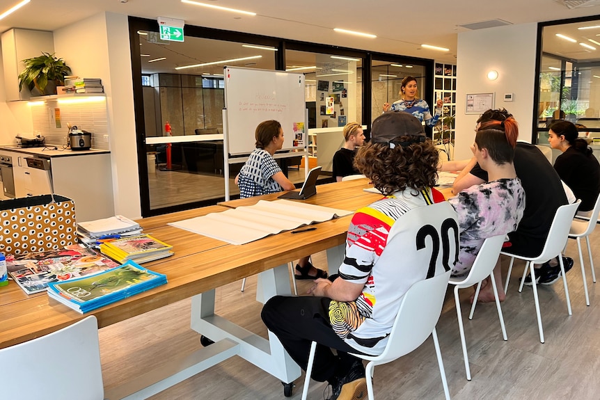 Six students sit at long wooden table, with teacher next to white board