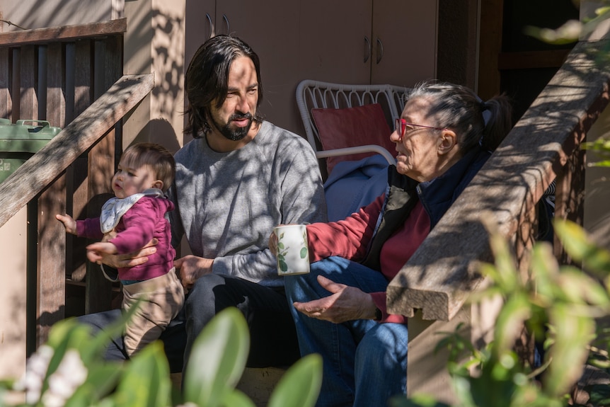 Luke and Sylvia sit on the back steps in the sunshine.  Sylvia holds a cup of tea and Luke holds his baby son on his lap from him. 