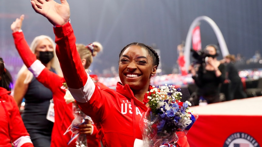 Gymnast simone biles waves to the crowd while smiling and holding flowers wearing a red team usa tracksuit