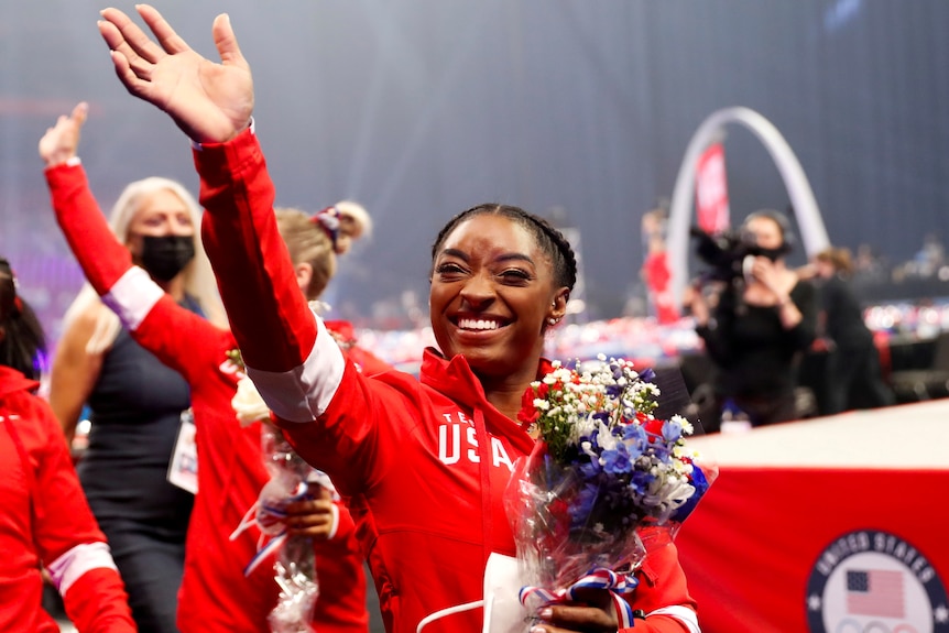 Gymnast simone biles waves to the crowd while smiling and holding flowers wearing a red team usa tracksuit