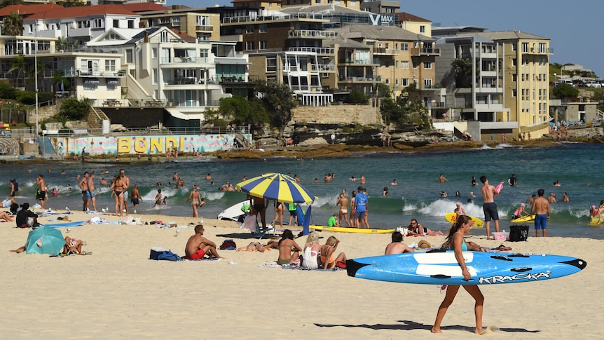 people sun baking, swimming and a woman carrying a surfboard at a beach