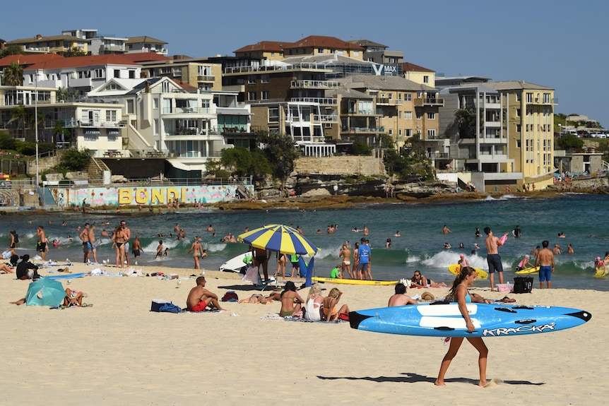 people sun baking, swimming and a woman carrying a surfboard at a beach