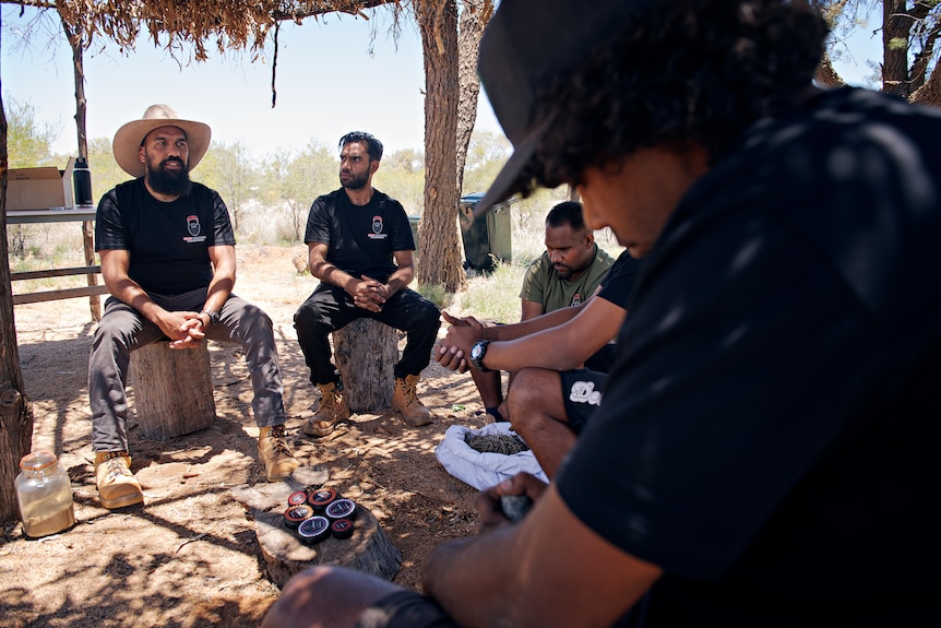 Men sit in a circle under a shelter in the bush.