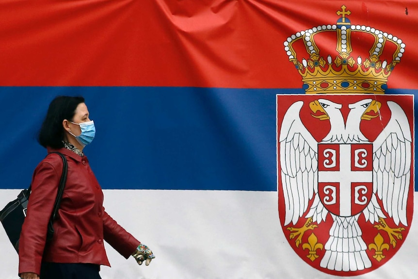 Una mujer con mascarilla y chaqueta roja pasa junto a la bandera serbia.