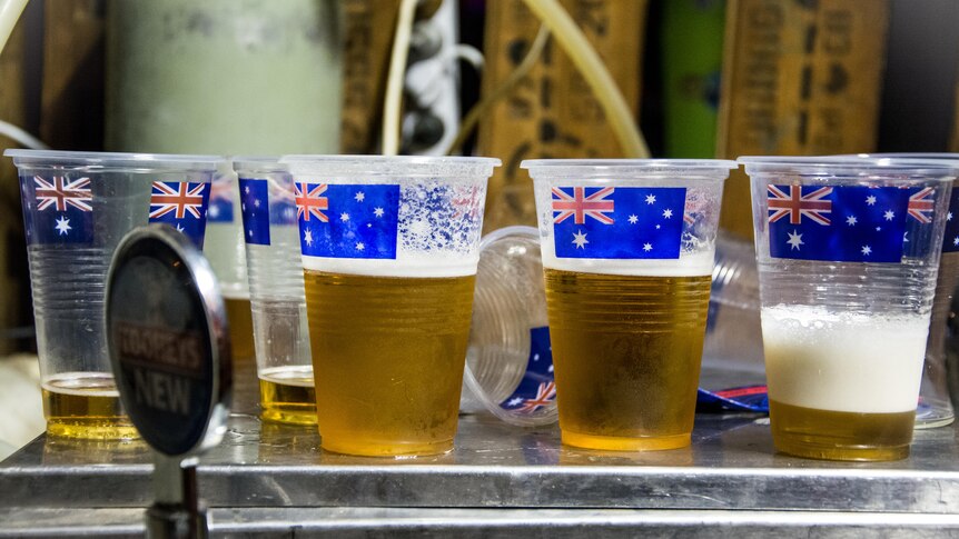 Four clear plastic cups of beer are lined up on a bar, each adorned with a picture of the Australian flag.