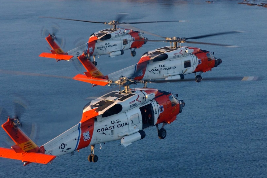 Three US Coast Guard helicopters fly over a stretch of dark blue water.