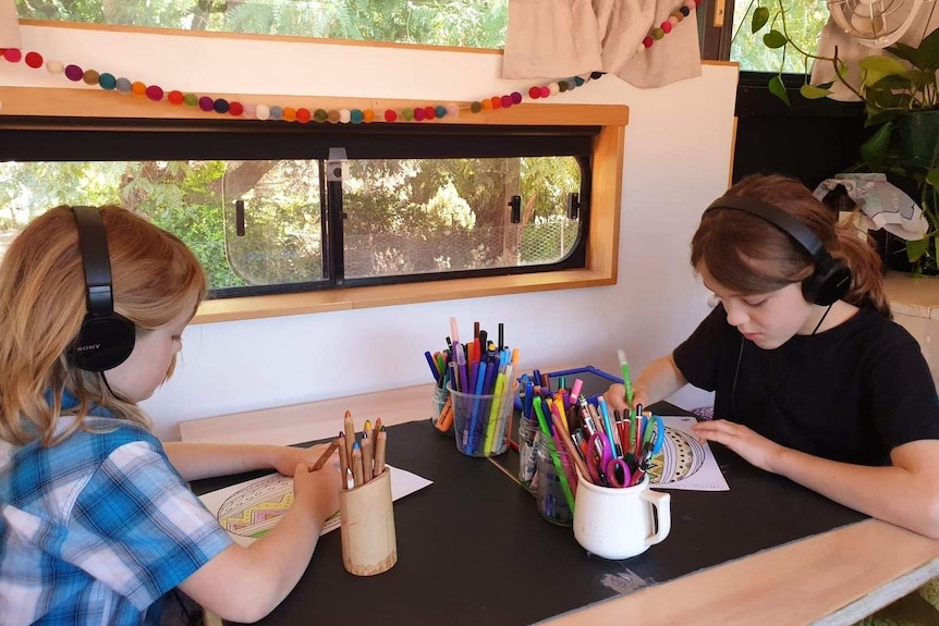 Ellery and Aubrey sits at a table with stationery and headphones doing home-schooling in their bus home.