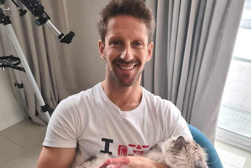 Romain Grosjean sits on a blue chair with a white t-shirt on, cuddling a fluffy cat in his lap