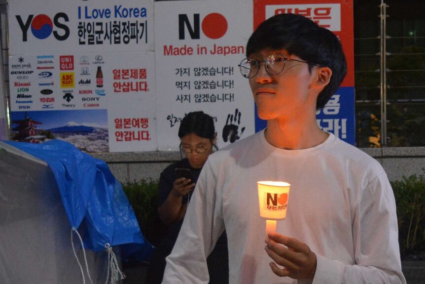 A man with round glasses stands holding a candle and looking into the distance in front of posters calling for a Japan boycott.