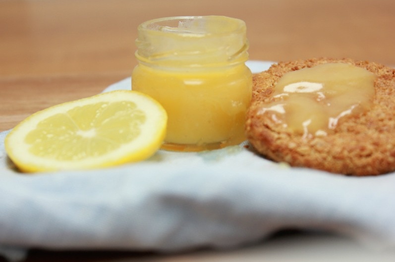 A small jar of lemon curd is open and sitting on a white cloth. There is a lemon slice on one side and a biscuit on the other.
