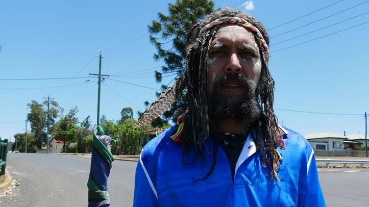 An Indigenous man with long hair, holding a long stick wrapped in colourful material, walks on a road under a blue sky.