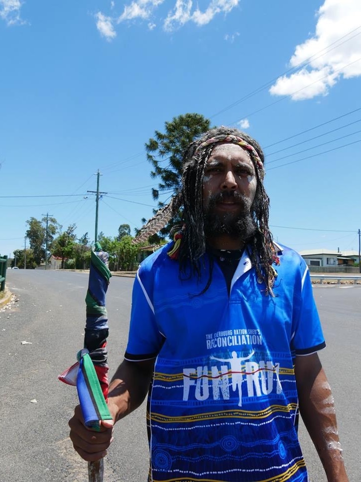 An Indigenous man with long hair, holding a long stick wrapped in colourful material, walks on a road under a blue sky.