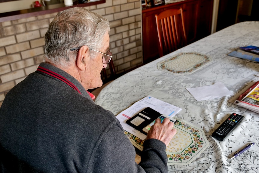 A man sitting at a table looking at a phone and telstra bills on the table. 