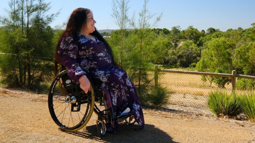 A woman in a pruple dress and a wheelchair smiles at a vista of green trees and rolling hills