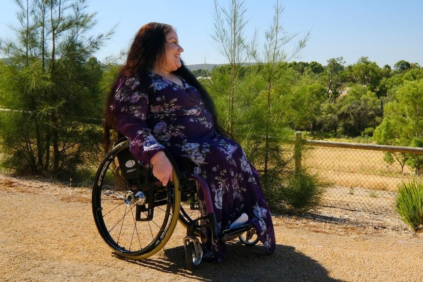 A woman in a pruple dress and a wheelchair smiles at a vista of green trees and rolling hills