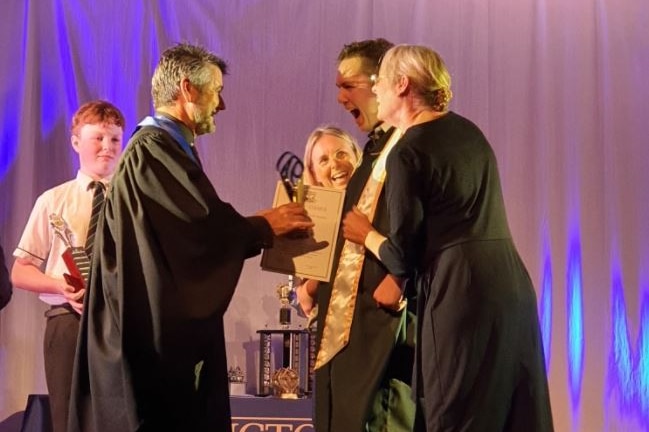 Man in academic robes hands trophy to Tristan Sik who is shouting with joy while standing with support worker