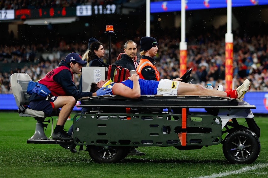 An AFL player lies on a stretcher and is carted off with support staff around him.