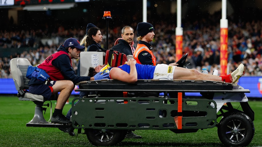 An AFL player lies on a stretcher and is carted off with support staff around him.