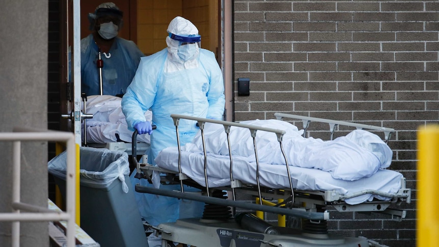 A person covered in protective gear wheels a body on a stretcher out of double doors. Behind, another is doing the same.