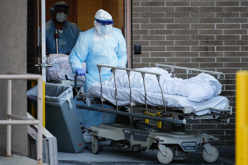 A person covered in protective gear wheels a body on a stretcher out of double doors. Behind, another is doing the same.