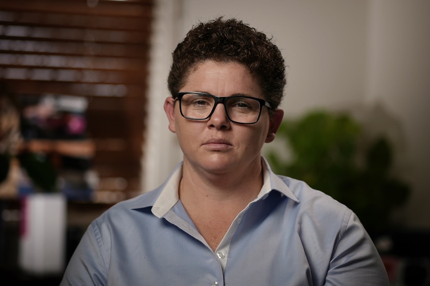 Woman wearing light blue shirt and glasses. She has short, brown, curly hair.