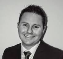 Shane Kuflik con traje sonríe a la cámara en una fotografía en blanco y negro. 