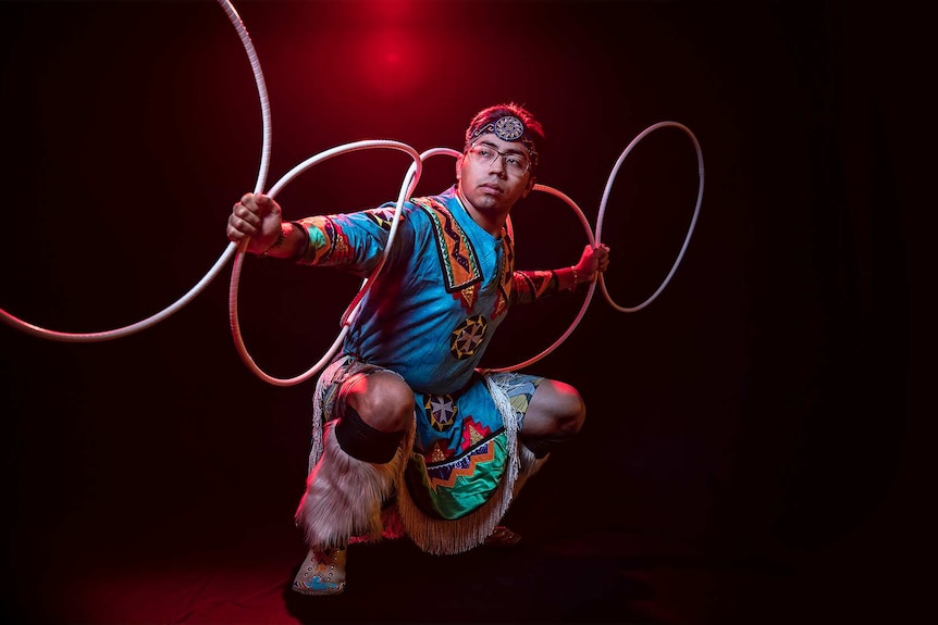 A man in bright blue Native American regalia poses in squat position with 5 hoops arranged across his back and outstretched arms