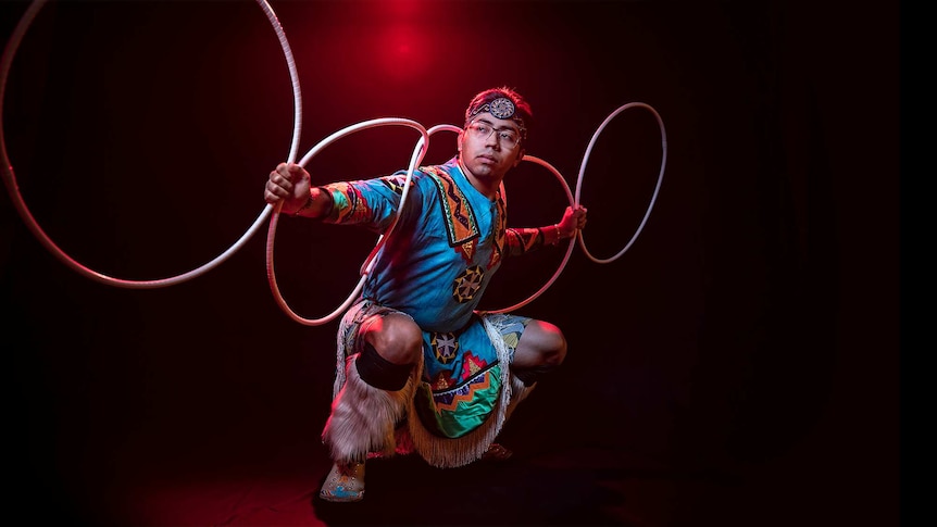 A man in bright blue Native American regalia poses in squat position with 5 hoops arranged across his back and outstretched arms