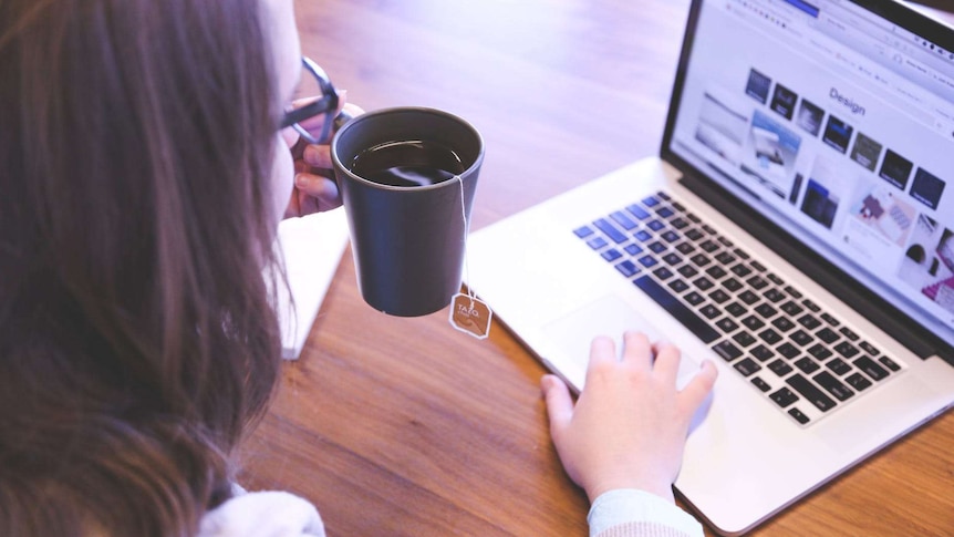 A woman sits at her kitchen table, drinking tea and using her laptop.