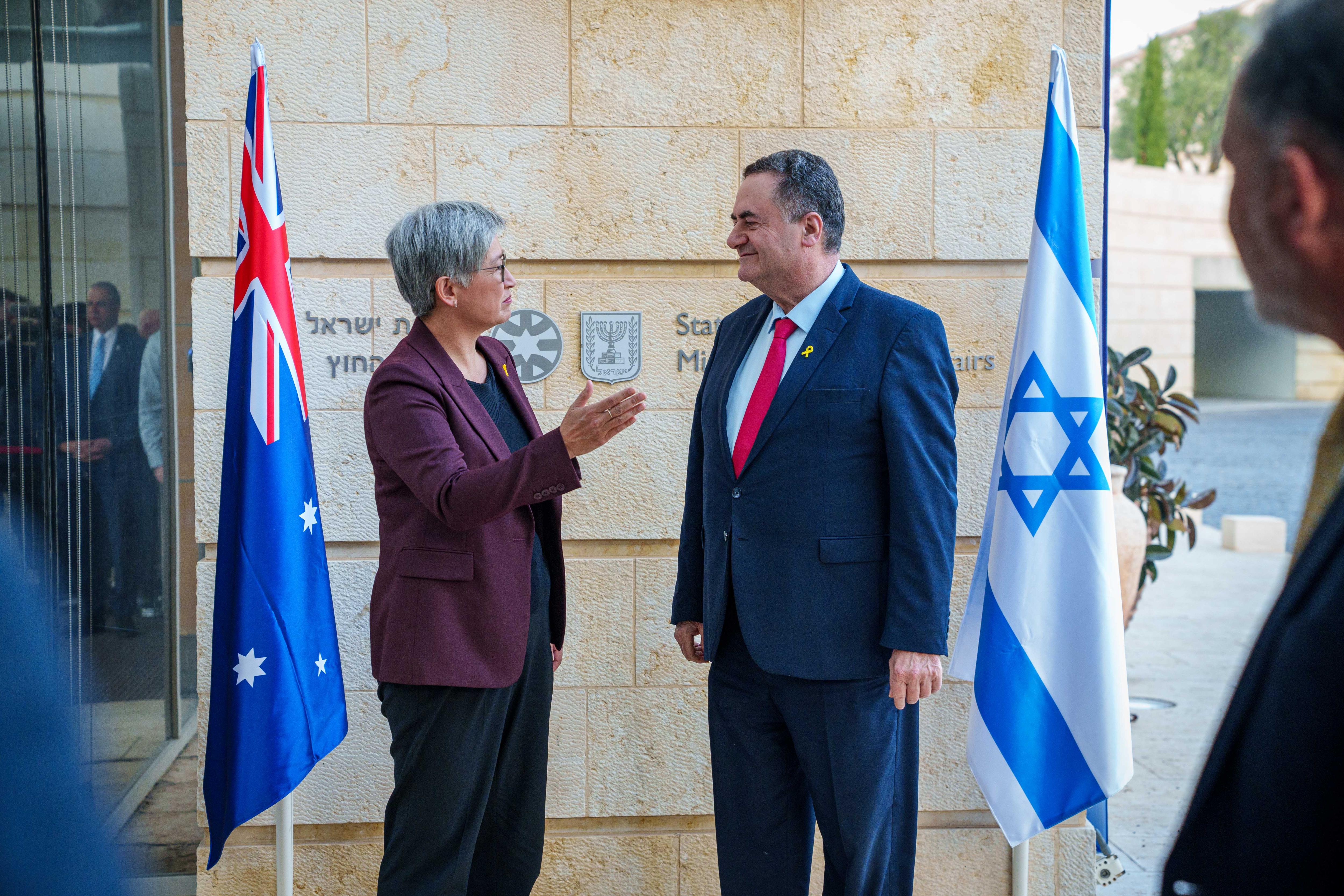 A man and woman, both in suits, stand and speak to each other in front of a building and Australian and Israeli flags.