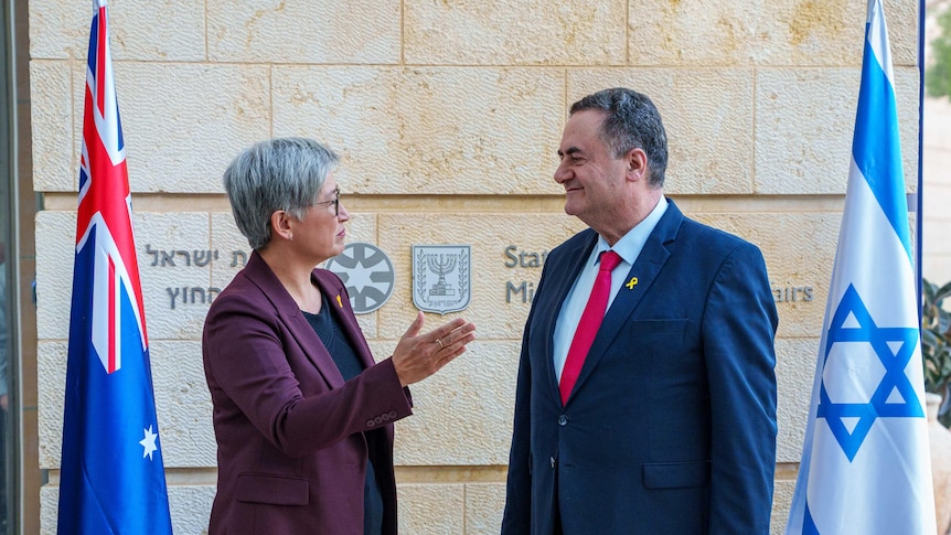 A man and woman, both in suits, stand and speak to each other in front of a building and Australian and Israeli flags.