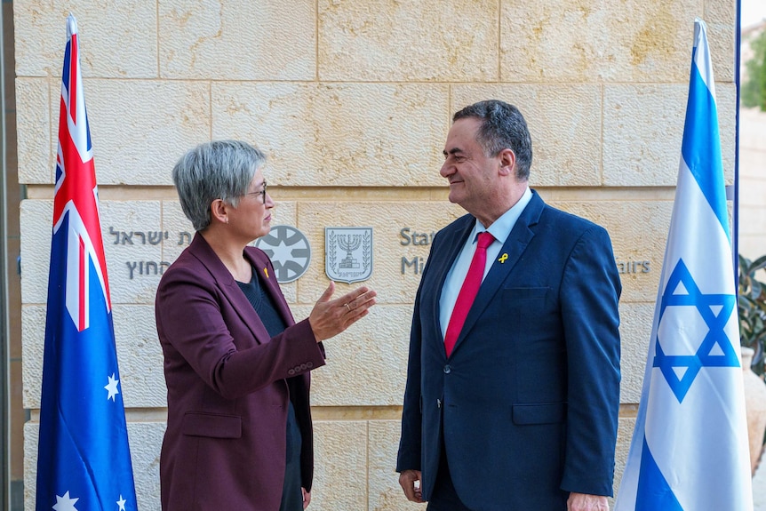Un bărbat și o femeie, îmbrăcați în costume, stau și vorbesc unul cu celălalt în fața unei clădiri și a steagurilor Australiei și Israelului.