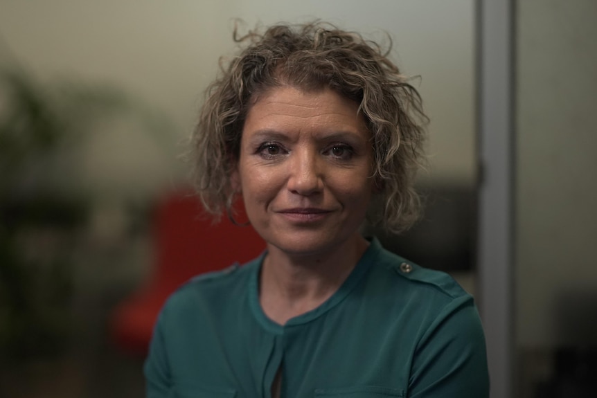 A woman with a green jumper and brown curly hair looks into the camera.