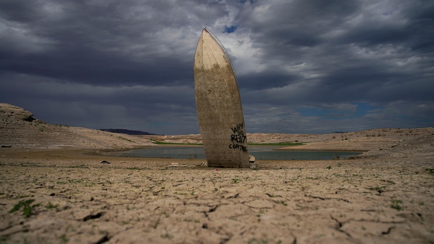 The hull of a boat stands directly upright in a lake bed. 3/4 of the boat are dark where it was previously submerged. 