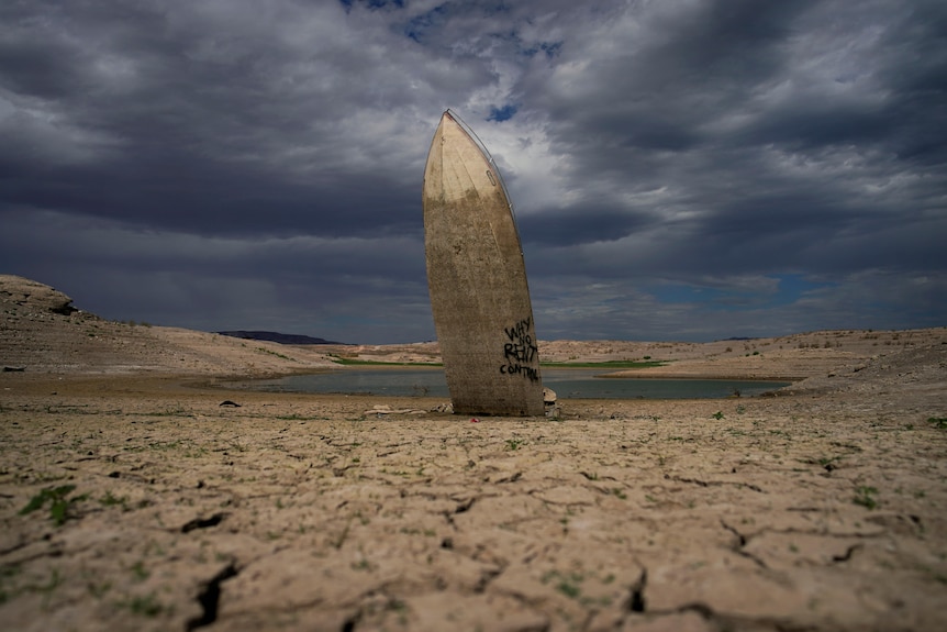 The hull of a boat stands directly upright in a lake bed. 3/4 of the boat are dark where it was previously submerged. 
