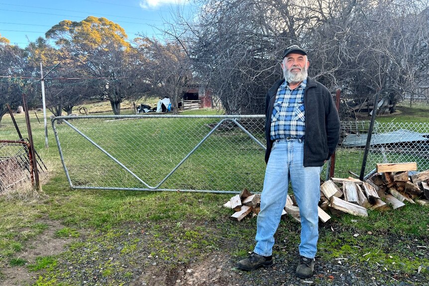 A man with a silver beard stands in front of a wire gate on a property.