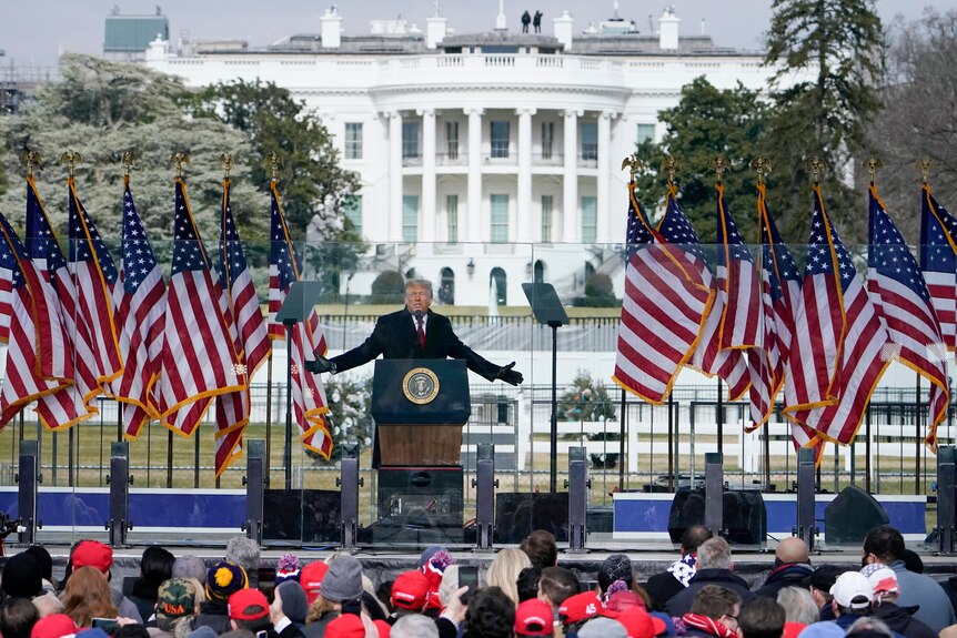 Donald Trump z rozpostartymi ramionami, stojąc na zewnętrznym podium przed Białym Domem 