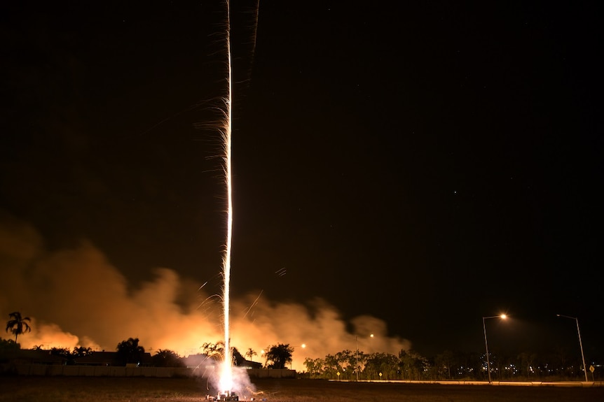 A firework goes off, as a small grassfire burns in the background