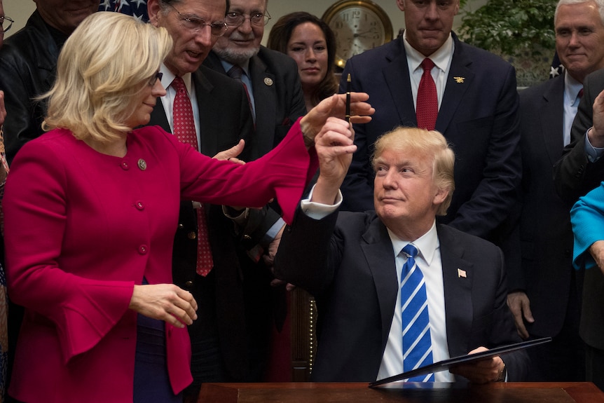 Donald Trump, a tight-lipped half-grin on his face, holds up a pen towards Liz Cheney. She wears a magenta jacket
