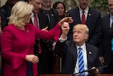 Donald Trump, a tight-lipped half-grin on his face, holds up a pen towards Liz Cheney. She wears a magenta jacket