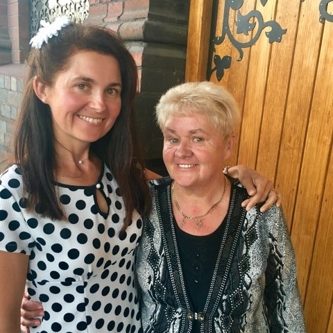 A woman aged in her 40s, wearing a spotted dress, stands next to her mother, with arms around each other.