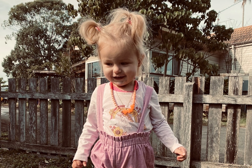A toddler with blonde hair in pigtails and pink overalls standing outside
