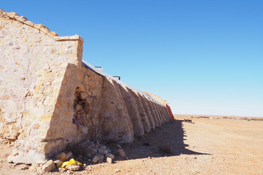 A large stone shed stands against a bright blue sky with buttressing in the foreground an an emergency orange tarp in the back.