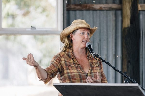 A woman in a hat and plaid shirt addresses a crowd from a microphone.