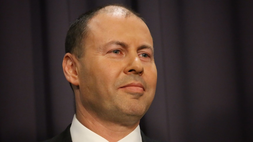 Headshot of Josh Frydenberg smiling against a dark background with a glimpse of a flag.