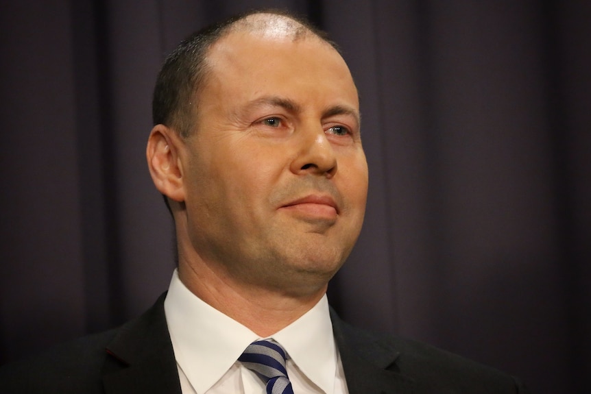 Headshot of Josh Frydenberg smiling against a dark background with a glimpse of a flag.