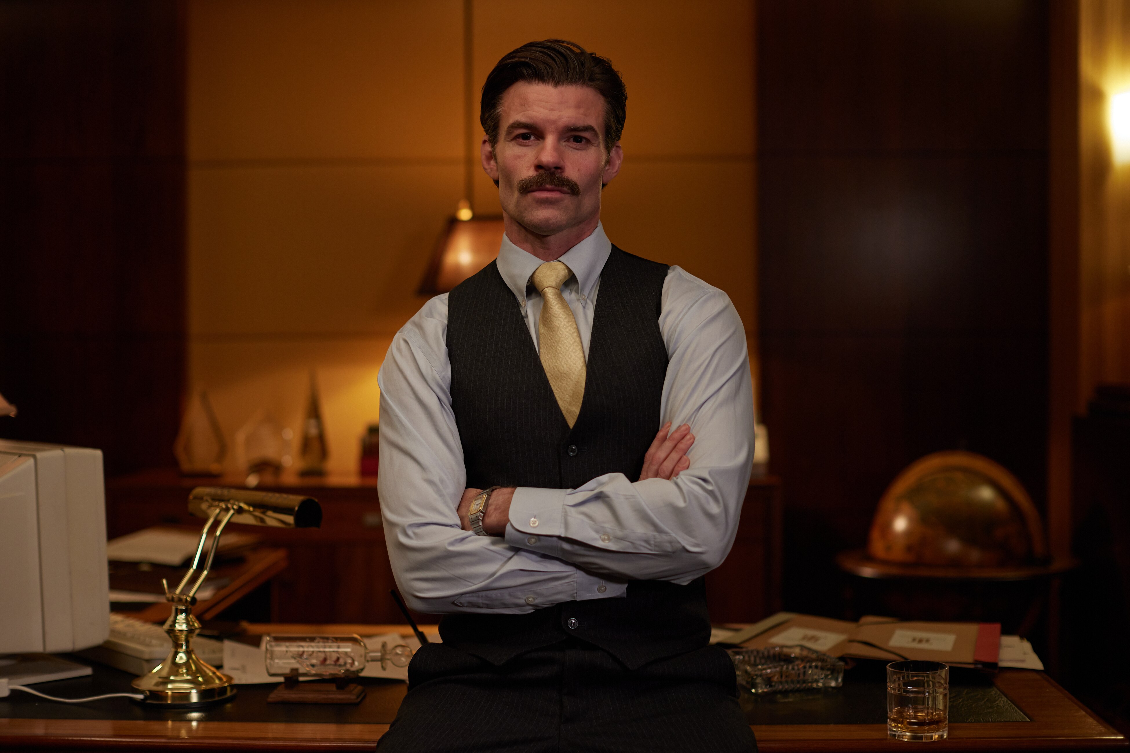 Daniel Gillies as CEO Charlie Tate in The Newsreader, sits on the front of his desk.