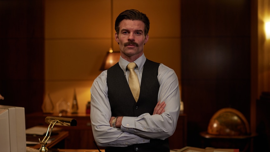 Daniel Gillies as CEO Charlie Tate in The Newsreader, sits on the front of his desk.